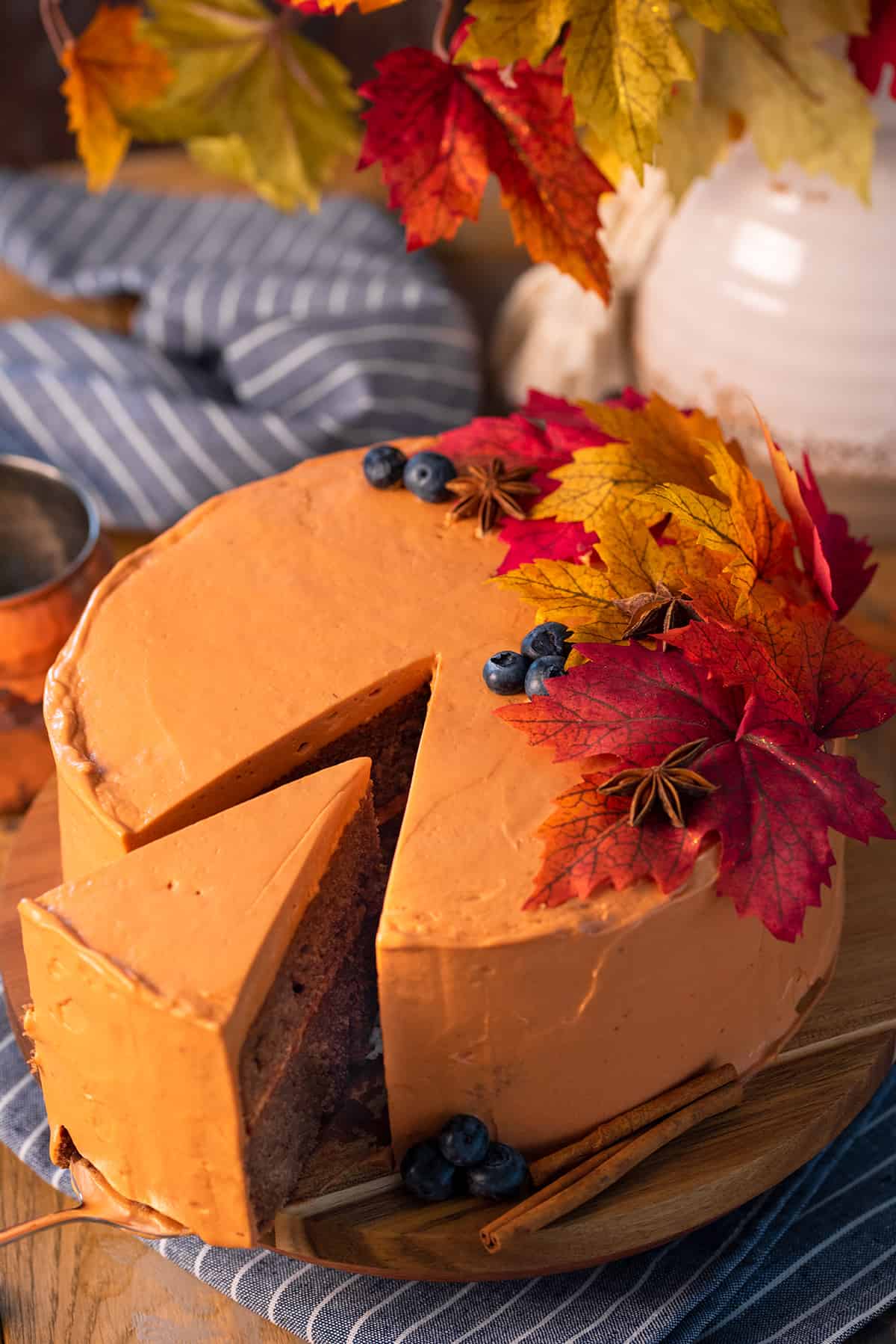 An orange fall cake decorated with red leaves and fall spices with a slice being pulled out of it.