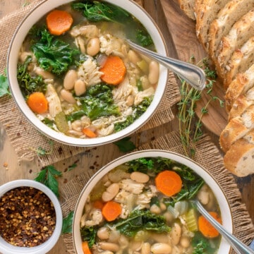 Two bowls of chicken soup with carrots, white beans, and kale in the soup and sliced bread in the background.