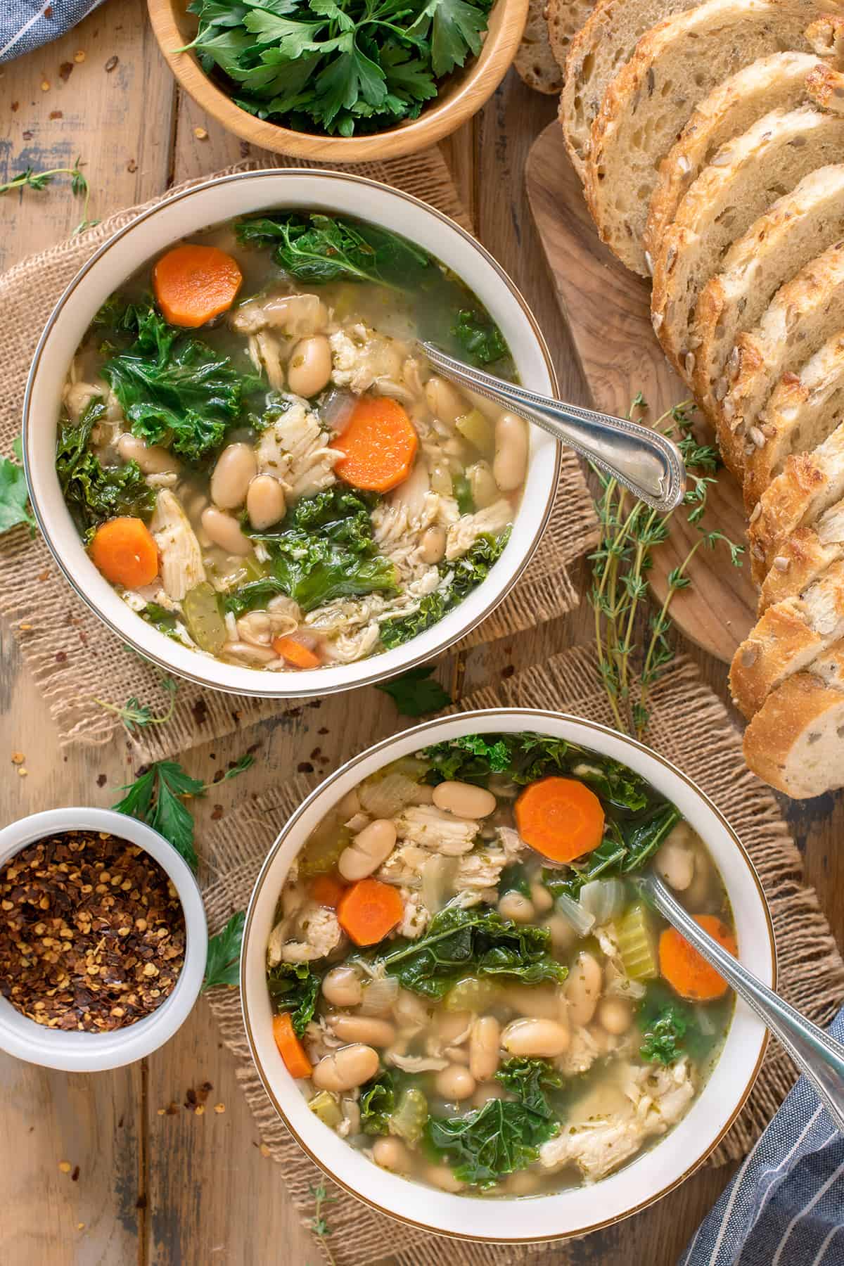 Two bowls of chicken soup with carrots, white beans, and kale in the soup and sliced bread in the background.