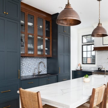 Behr Midnight blue painted cabinets in a classic old world kitchen with modern white accents and backsplash. Wicker chairs, copper light fixtures, and wood floors add warmth.