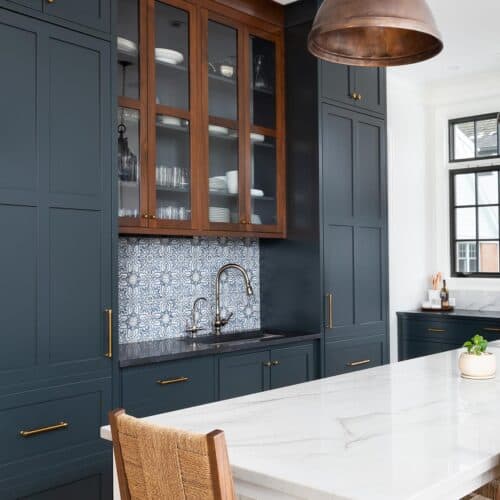 Behr Midnight blue painted cabinets in a classic old world kitchen with modern white accents and backsplash. Wicker chairs, copper light fixtures, and wood floors add warmth.