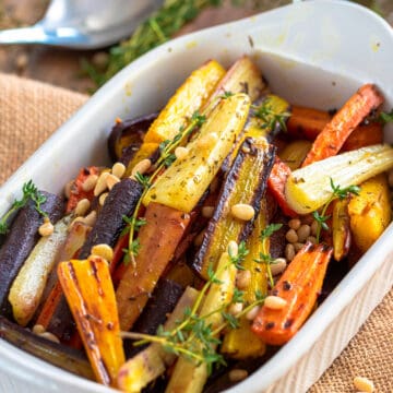 Roasted carrots in a white serving dish with fresh thyme and pine nuts sprinkled over top.