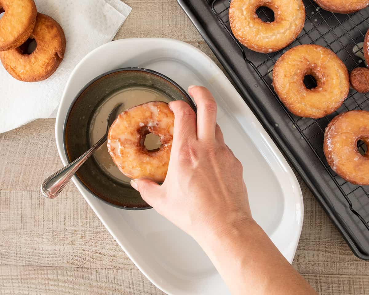 Overhead of hand glazing a donut with a hot water bath and doughnuts on the side cooling.