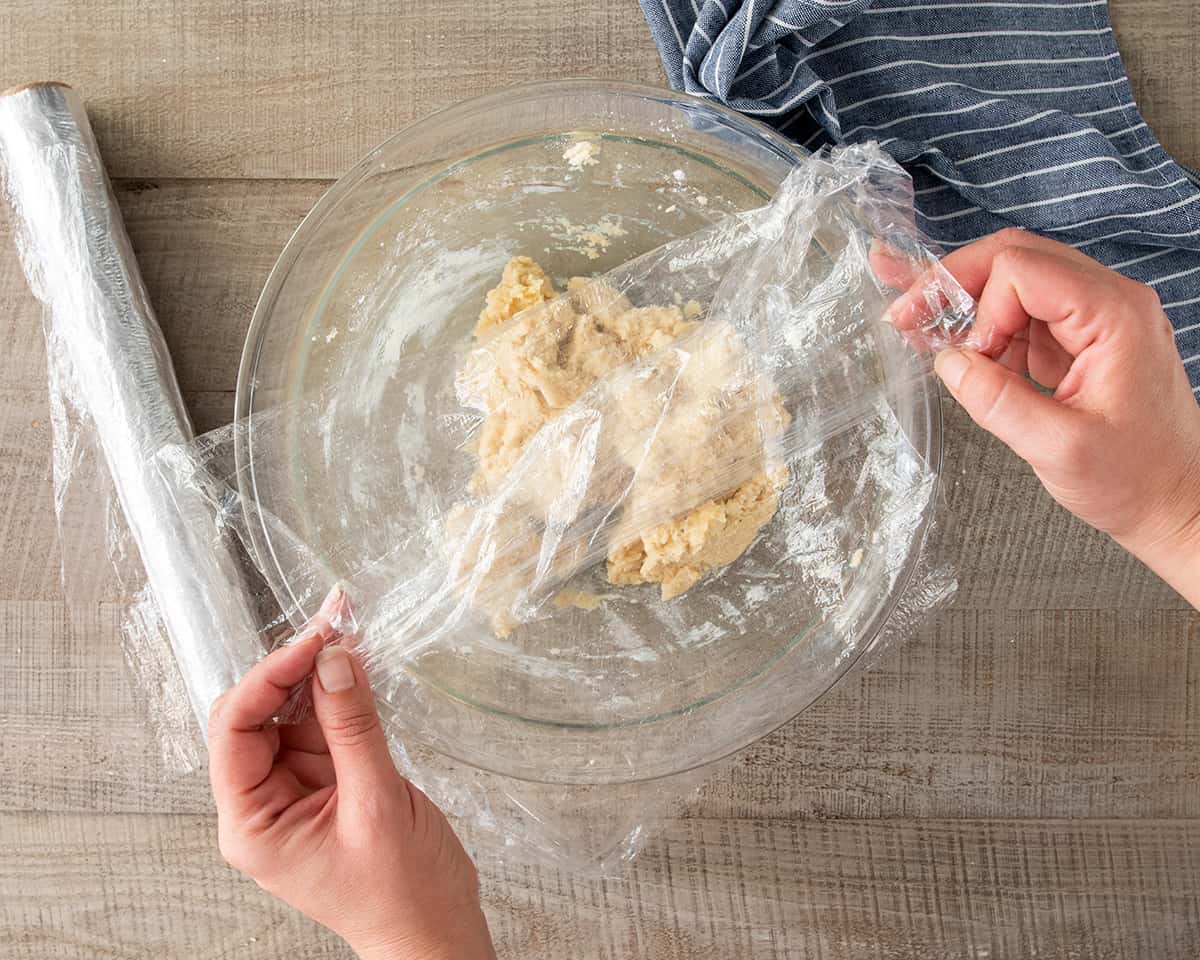 Dough for making donuts in a glass bowl with plastic wrap over it.