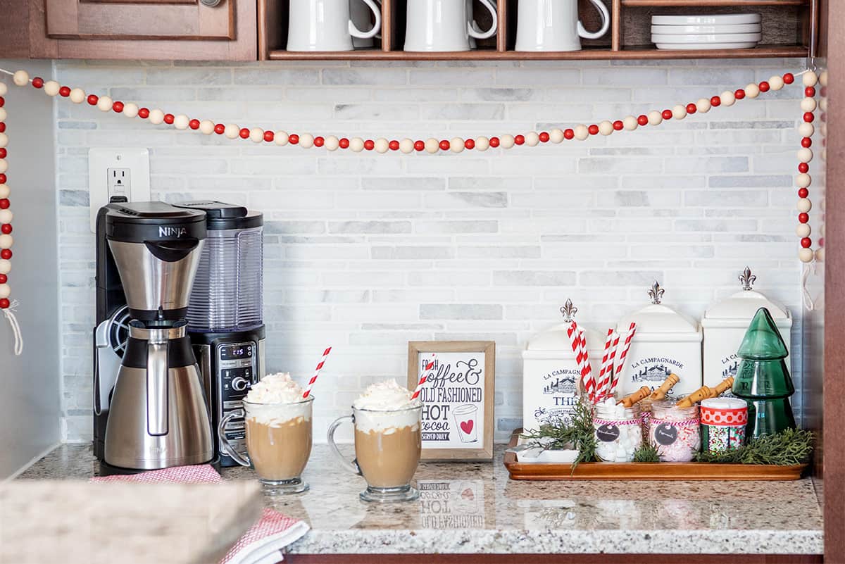 A simple holiday hot cocoa bar with coffee machine and jars of toppings on a serving tray with two cups of hot chocolate on the counter.