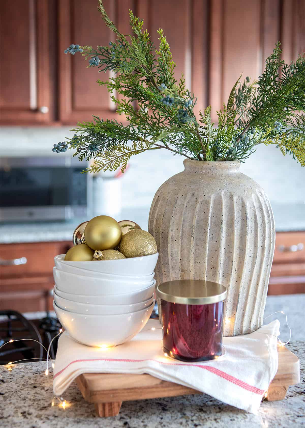 Christmas centerpiece on kitchen island with bowls, ornaments, candle, and vase of cedar branches.