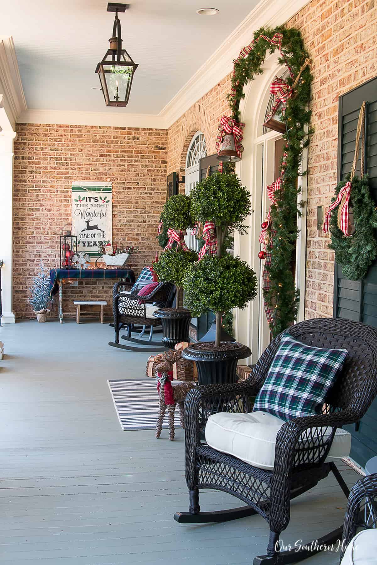 Large, covered porch Christmas tree topiaries in front of door with bells and bows around.