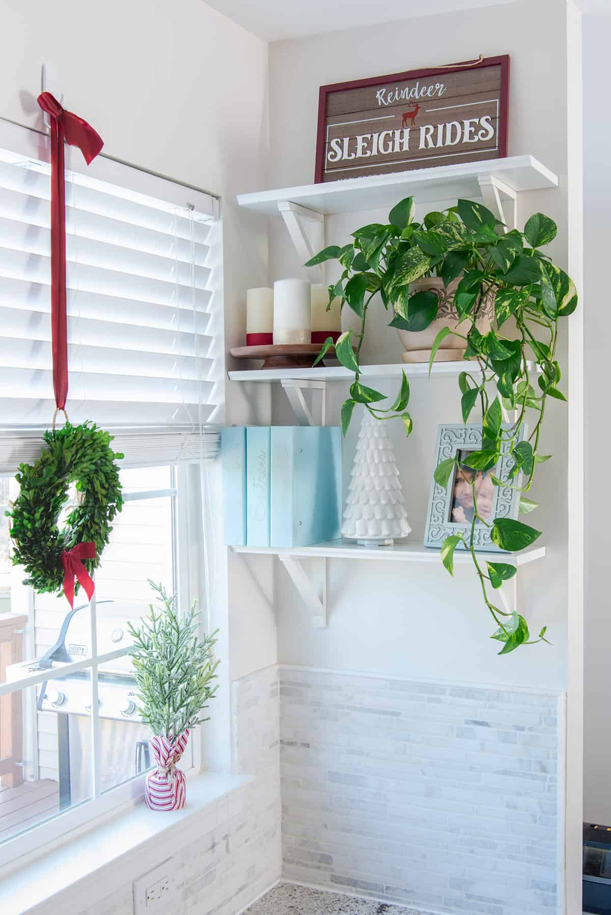 Christmas decorated open kitchen shelves with candles, wreath, and reindeer sign, and large houseplant.