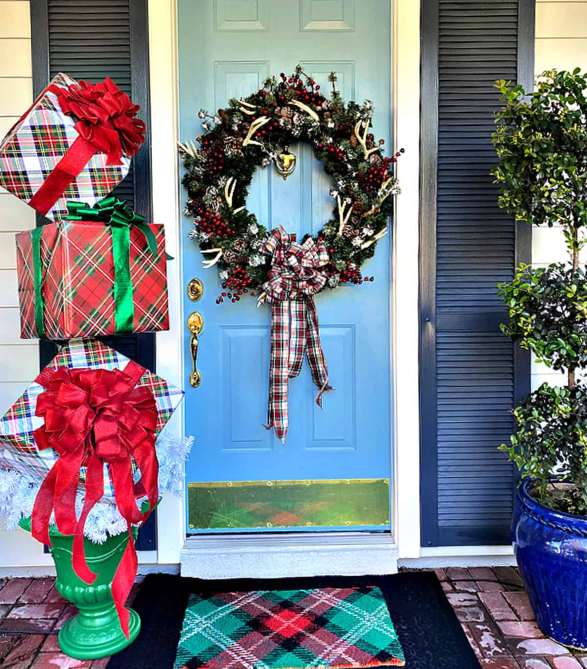 Stacked packages topiary for front porch Christmas decorations and a plaid wreath on blue door.