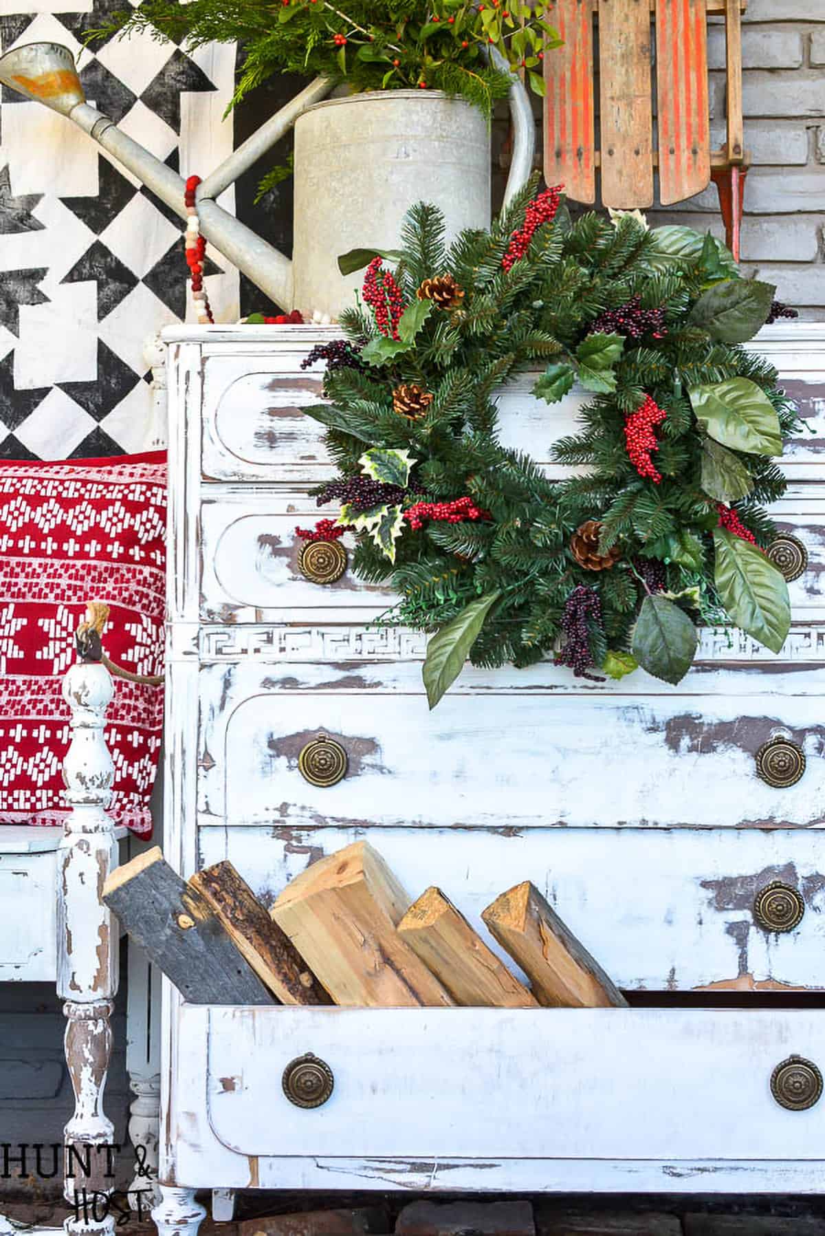 A distressed white dresser outside with a wreath on it and firewood in one drawer.