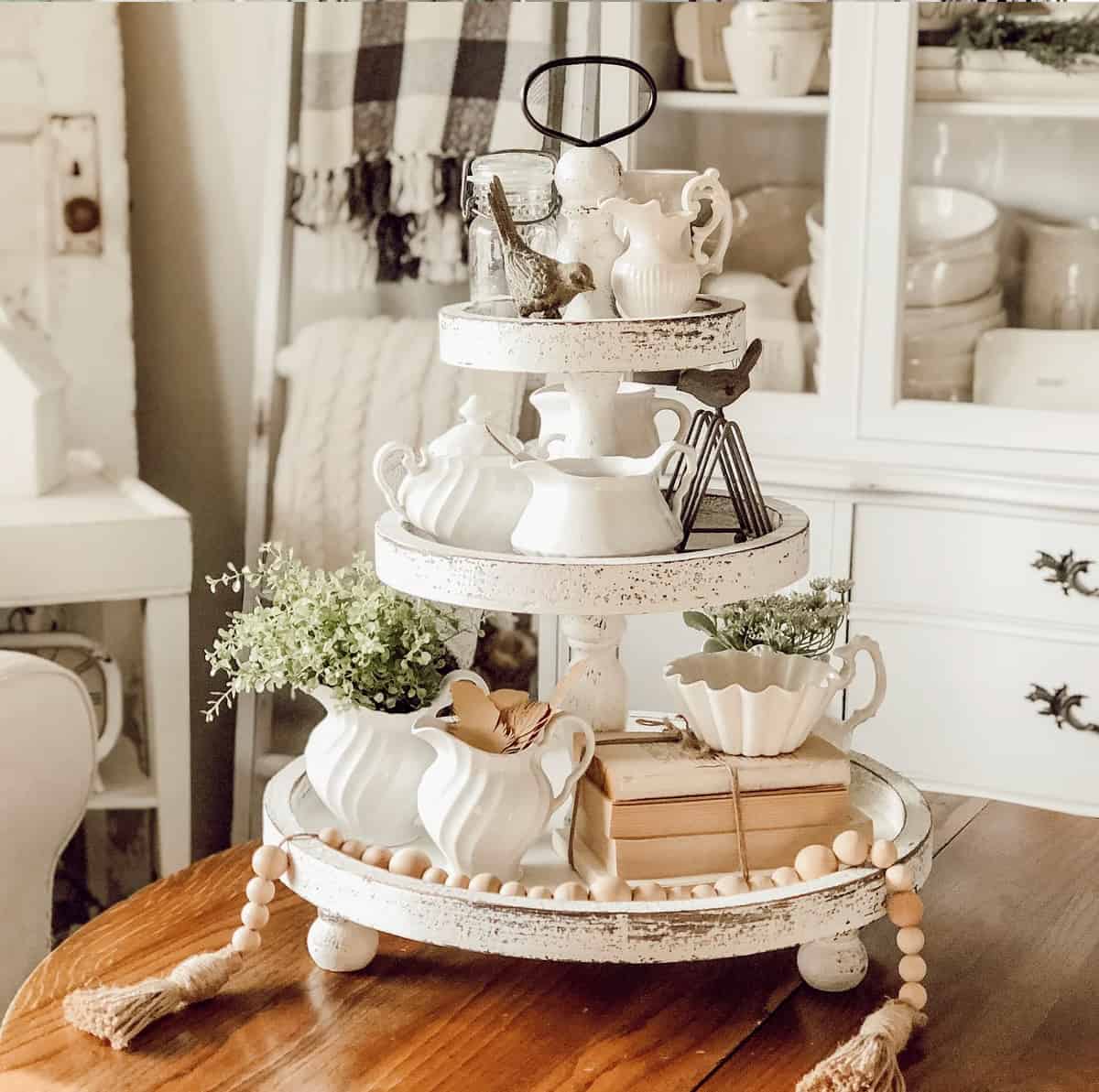 3-tiered distressed tray with farmhouse styled accessories and classic white creamery dishes surrounded by pewter birds, antique books, and fresh flowers.