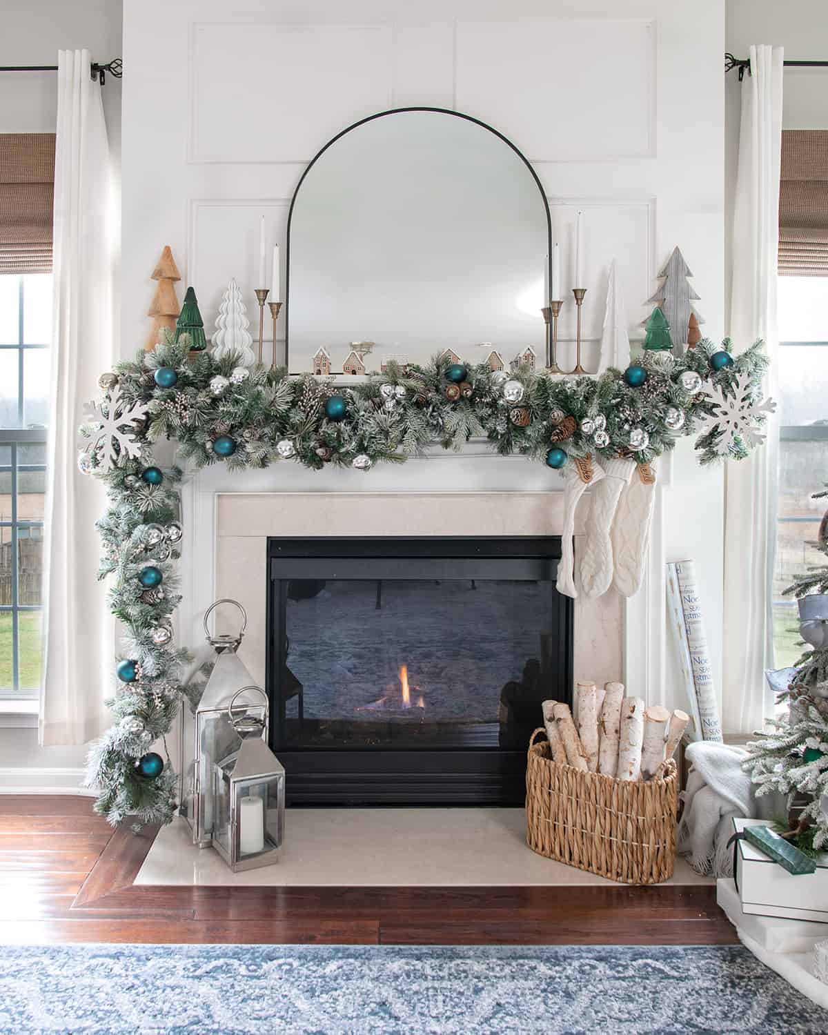 Green and silver Christmas mantel with neutral decorations, winter trees, and a large arched mirror over the fireplace.