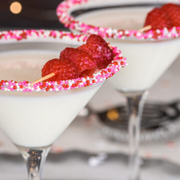 Two white chocolate martini glasses garnished with Valentine's Day sprinkles and raspberries.