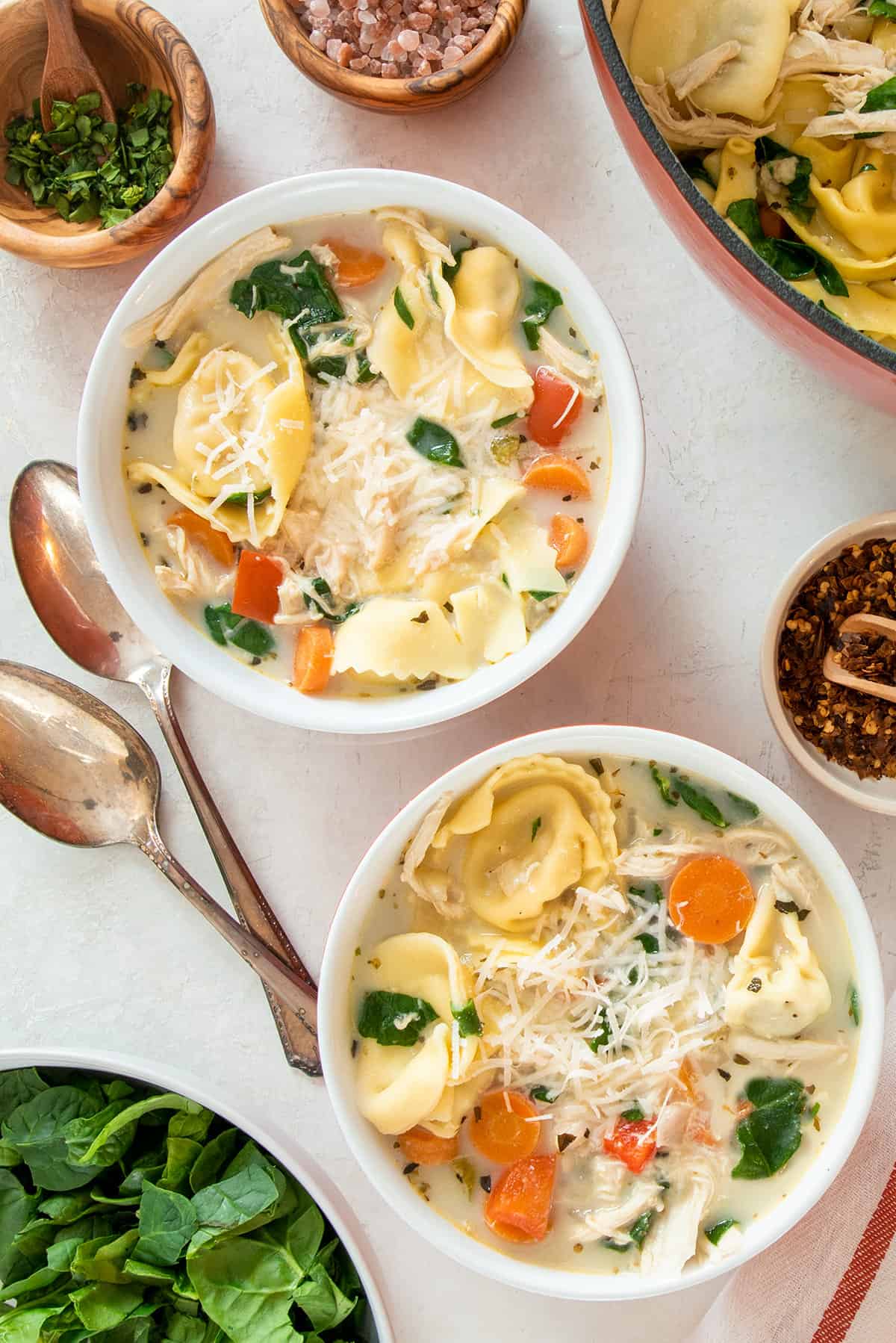 Overhead of chicken tortellini soup with spinach, carrots, and red peppers in two bowls with spoons on the side.
