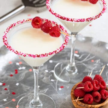 Two White Chocolate martinis with raspberry garnish on a silver tray for Valentine's day with a bowl of garnishes on the side and confetti.