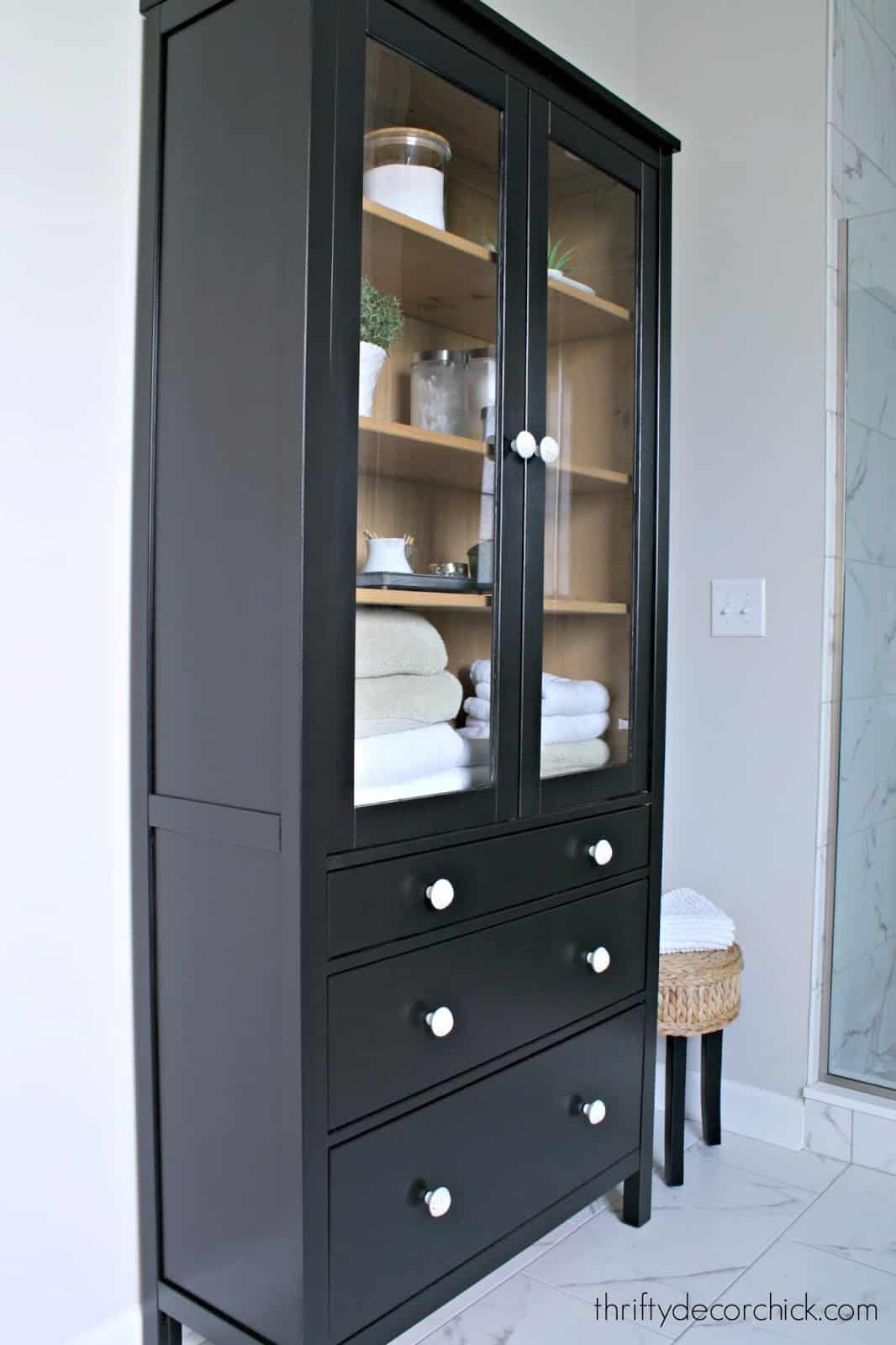 Glass door bathroom IKEA hutch painted black with linens and toiletries inside.