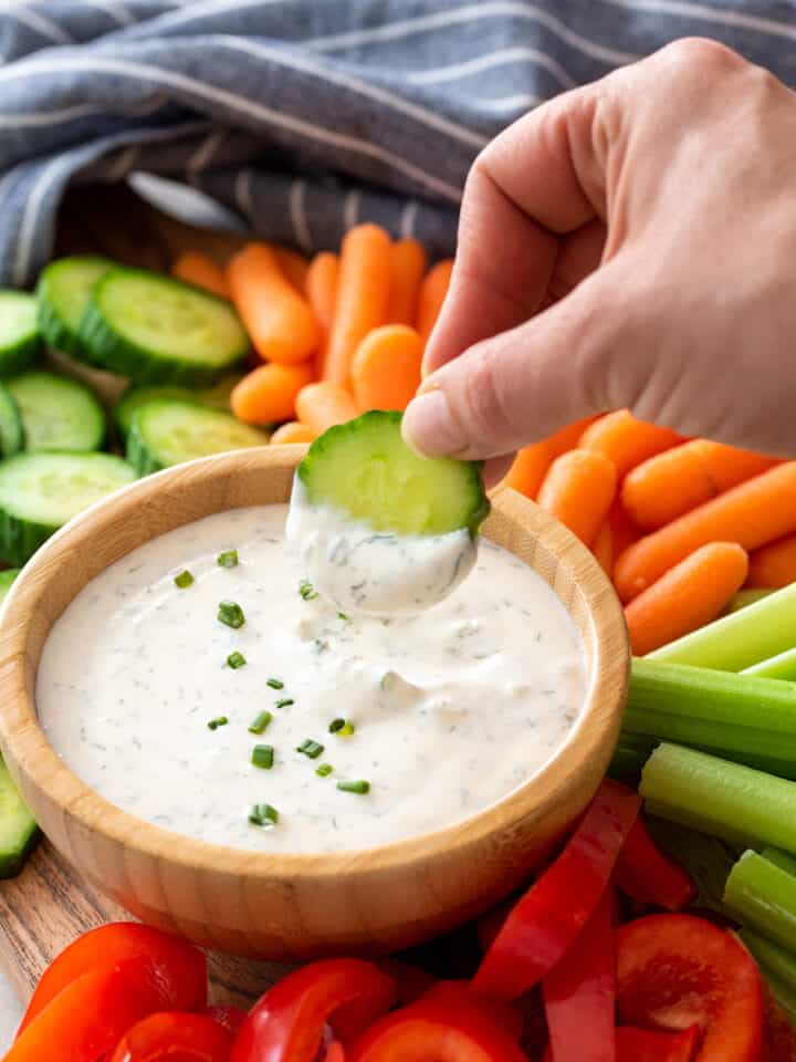 A hand dipping a cucumber into healthy ranch dip on a crudité platter.