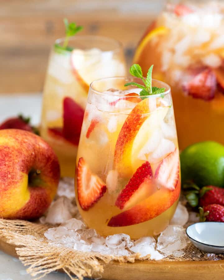 Two glasses of Moscato sangria with limes, apples, and peaches scattered around a serving tray.