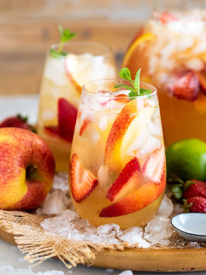 Two glasses of Moscato sangria with limes, apples, and peaches scattered around a serving tray.