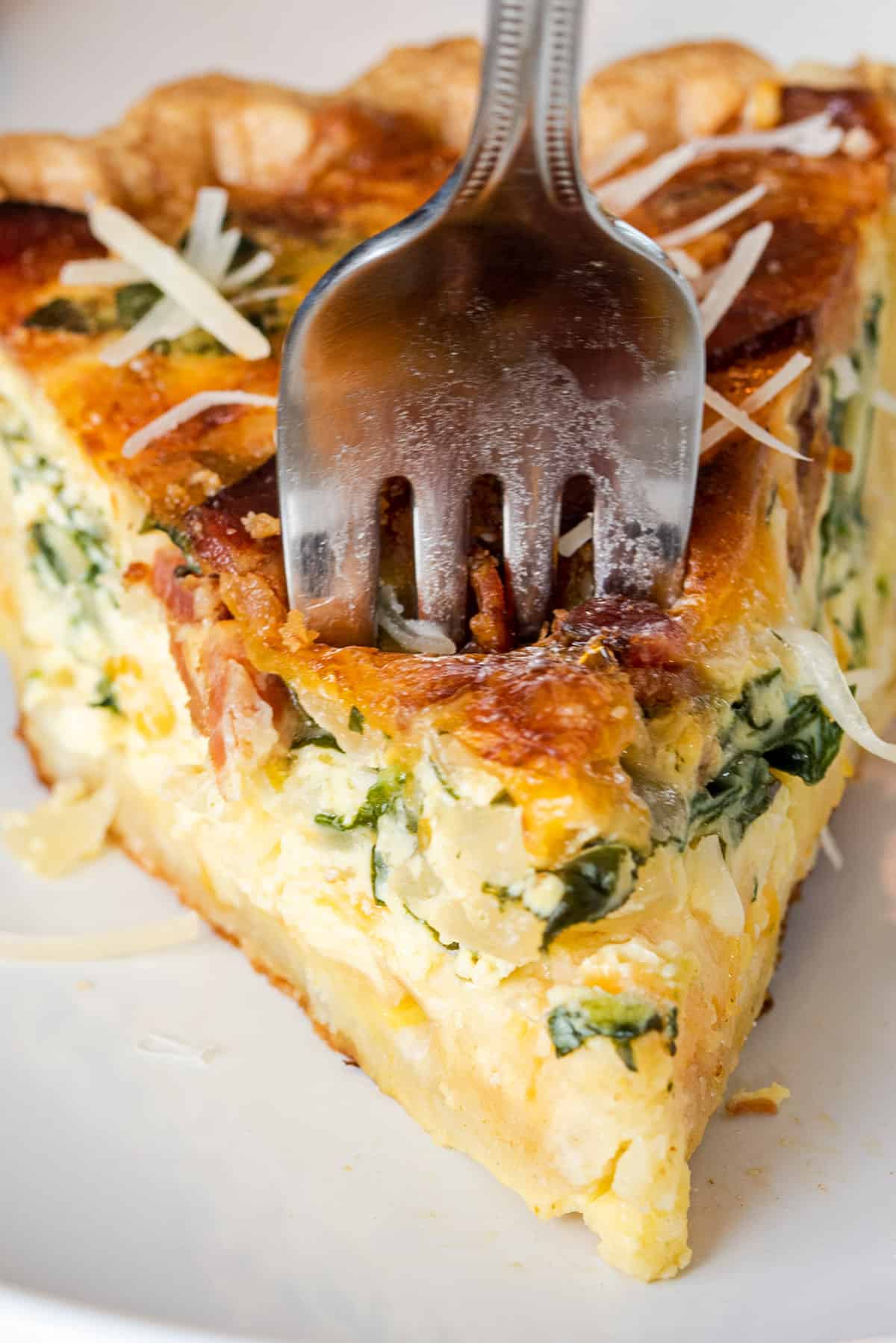 Slice of spinach bacon quiche with a fork stabbing through the top.