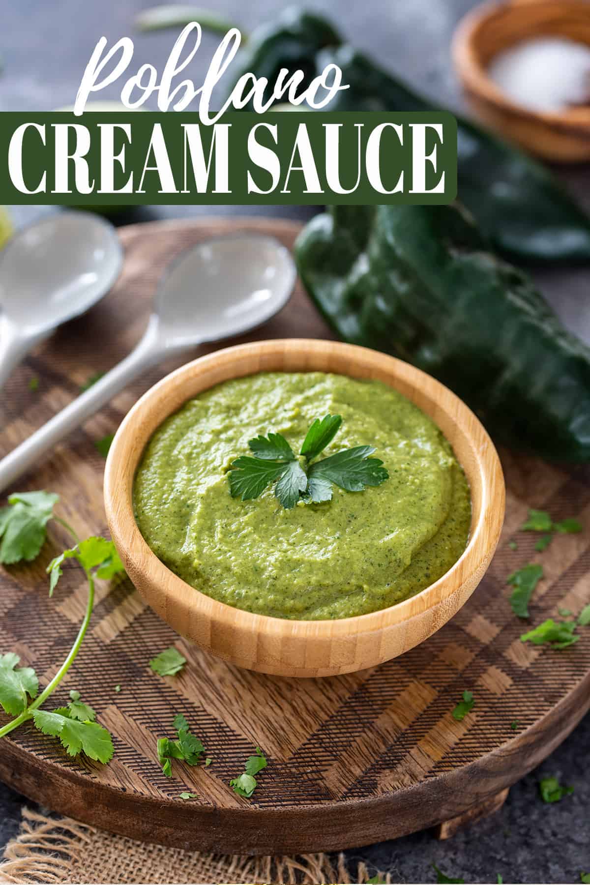 Bowl of green sauce with cilantro on top and peppers in the background on a plaid wood background.