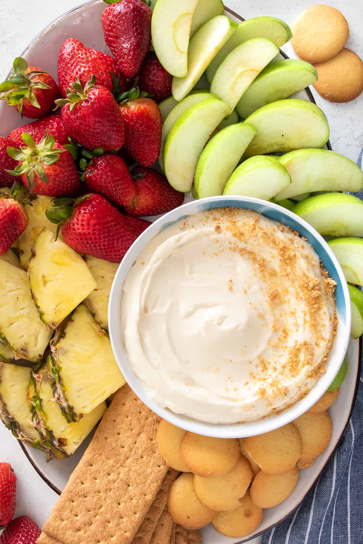 A bowl of cheesecake dip sprinkled with graham cracker crumbs and surrounded by cut fruit.
