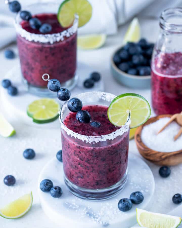 Two glasses of frozen blueberry margaritas with limes and blueberries sprinkled around a white background.