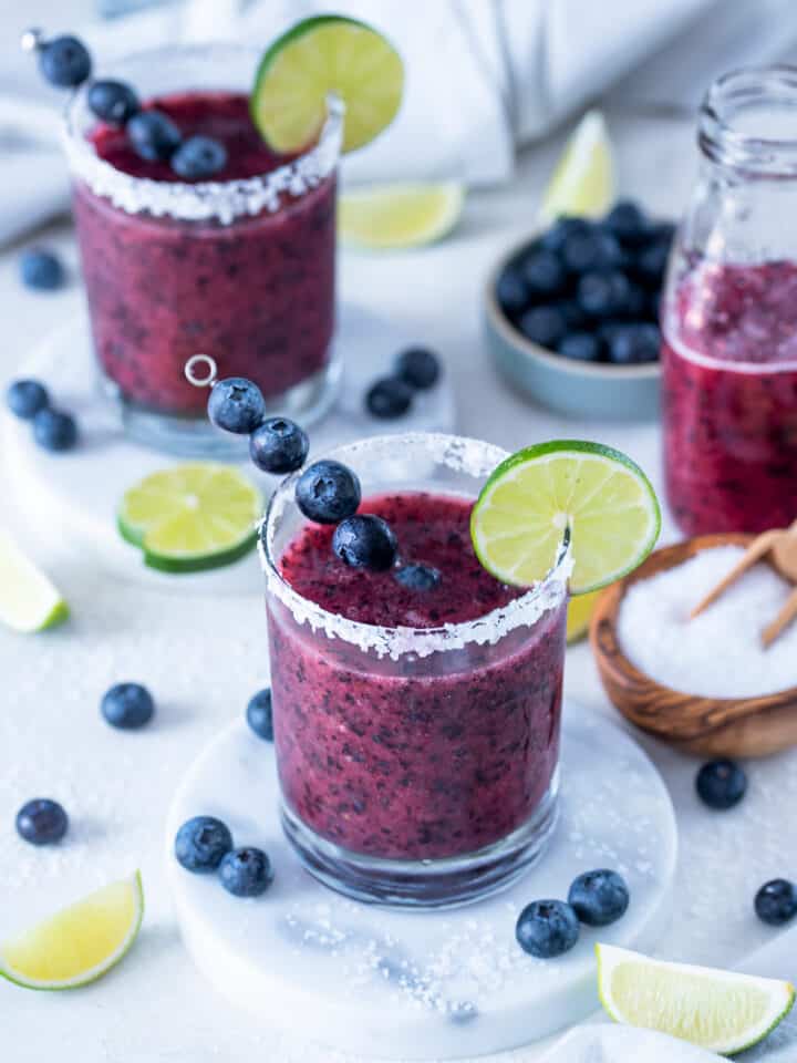 Two glasses of frozen blueberry margaritas with limes and blueberries sprinkled around a white background.