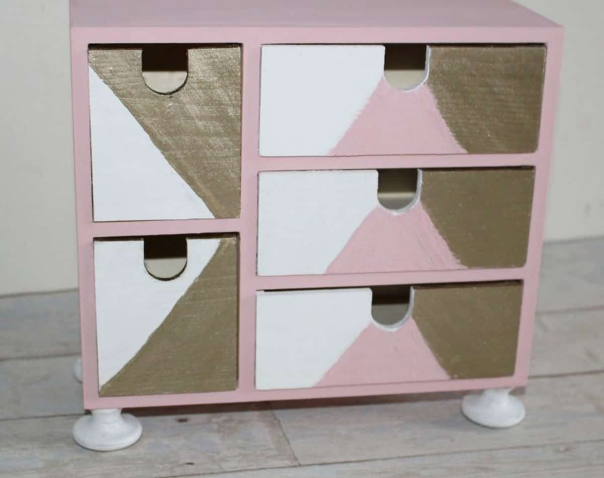 Ikea desk organizer painted in shades of pink, white, and gold with drawer knobs as feet.
