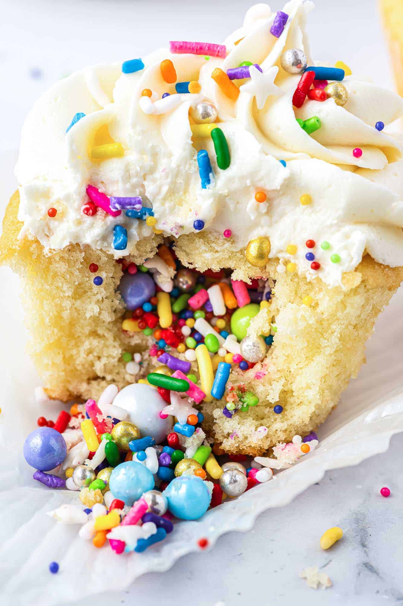 Pinata style vanilla cupcake with vanilla buttercream frosting opened to reveal cascade of rainbow sprinkles and topped with rainbow sprinkles.