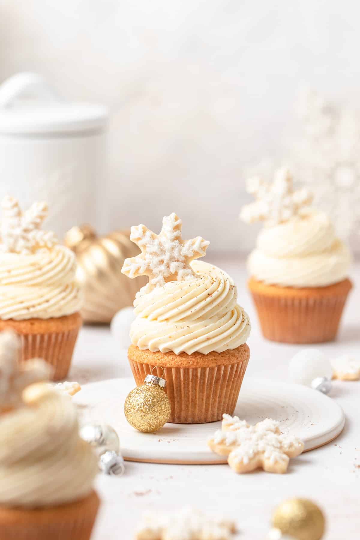 Eggnog cupcakes with white eggnog buttercream frosting and white snowflake cookie garnish with mini Christmas balls and baubles on white tabletop.
