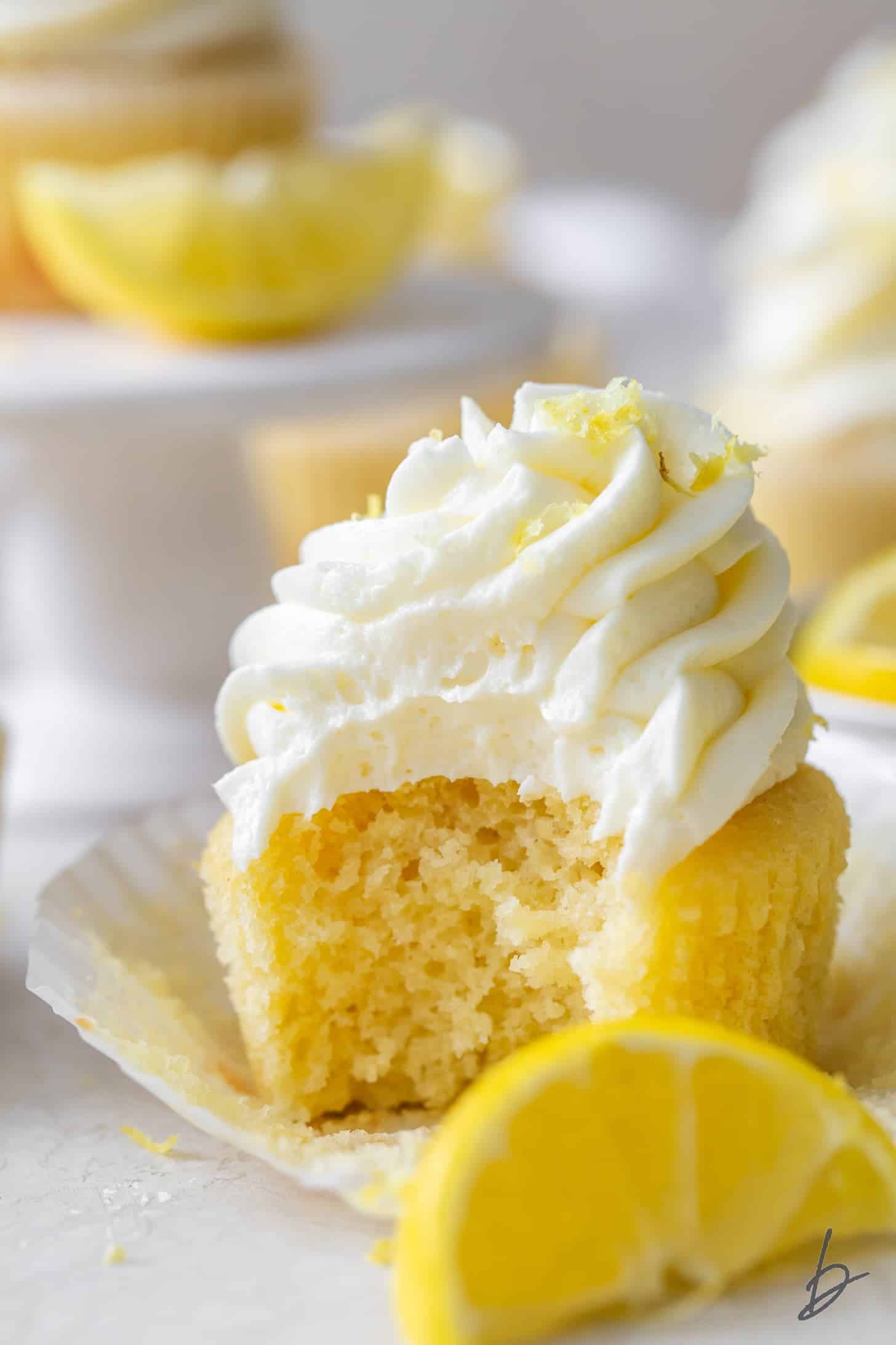 Lemon cupcake with white lemon buttercream with missing bite featuring lemon slices in back/foreground.