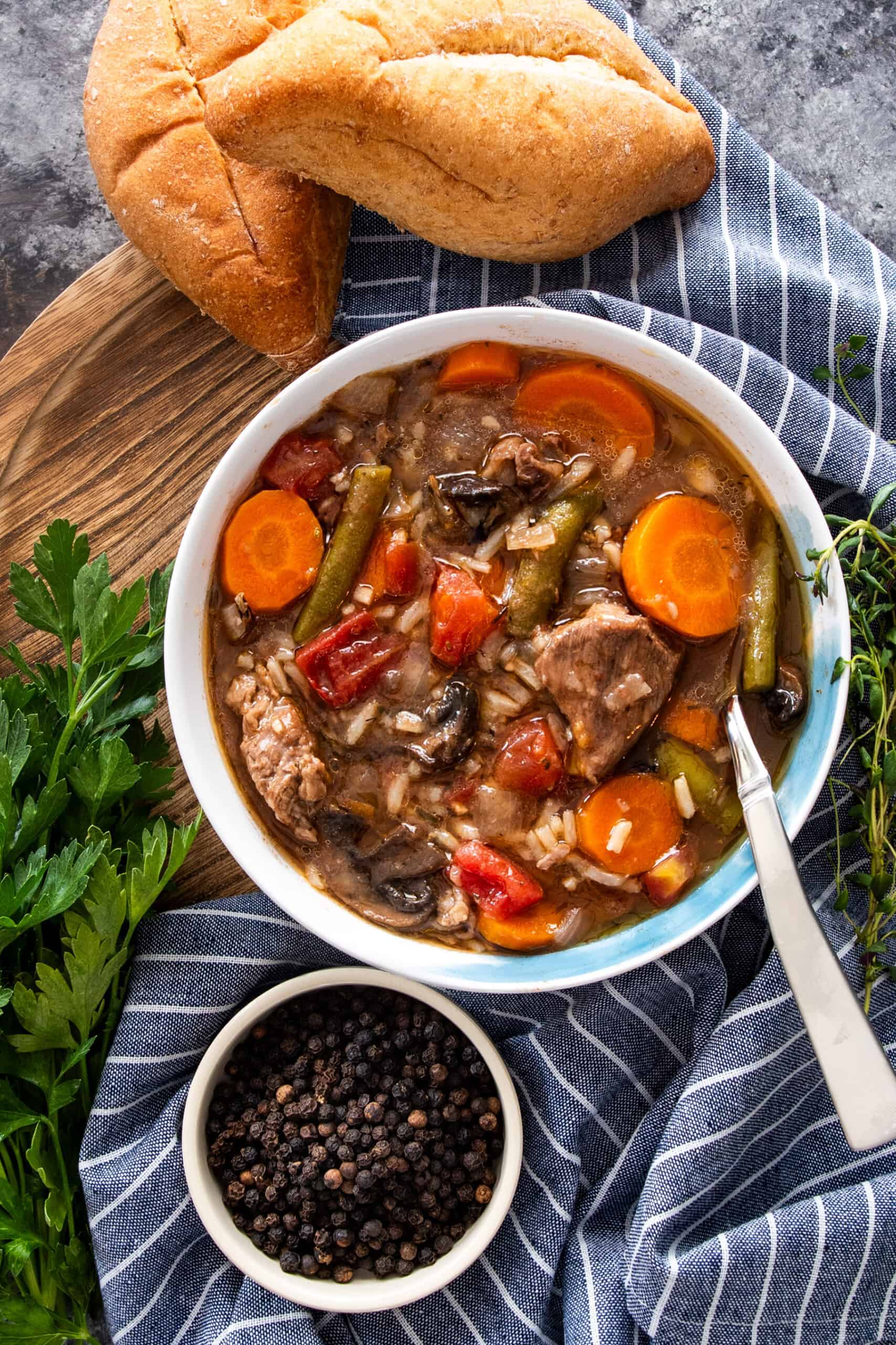 A bowl of beef and wild rice soup with black peppercorns on the side and a blue towel.