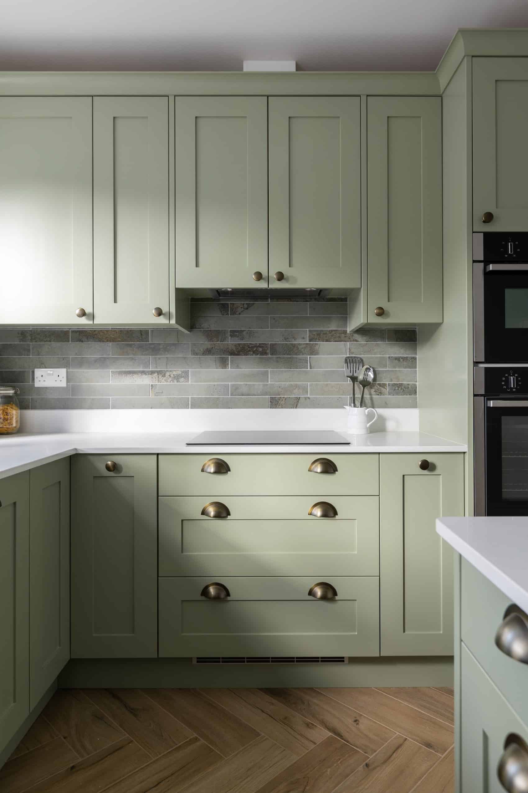 Closeup of green kitchen cabinets with antique brass pulls and white countertops.