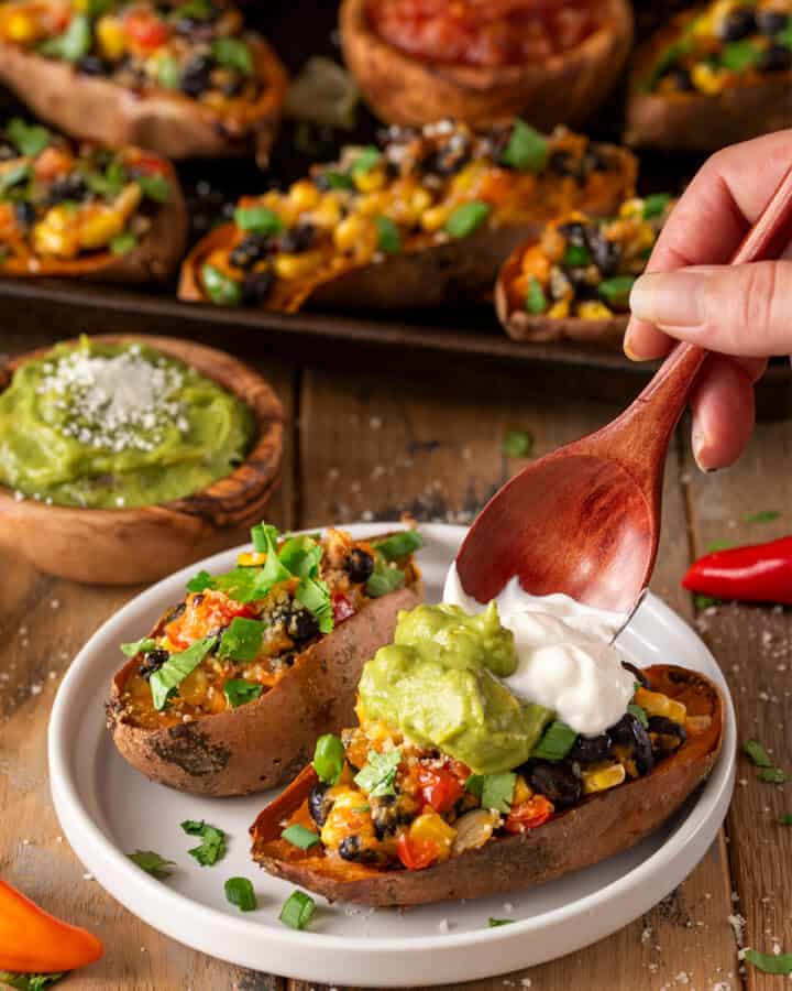 Two stuffed Mexican sweet potatoes in front while a hand tops them with guacamole and sour cream.