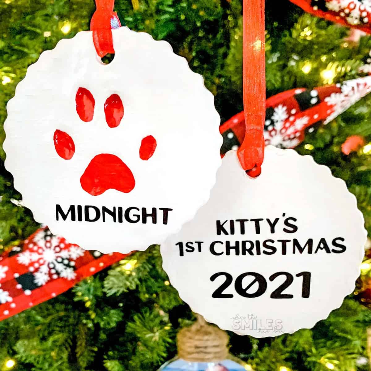 White clay Christmas tree ornaments hung with red ribbon- one with red paw print and both with black etching commemorating pets.