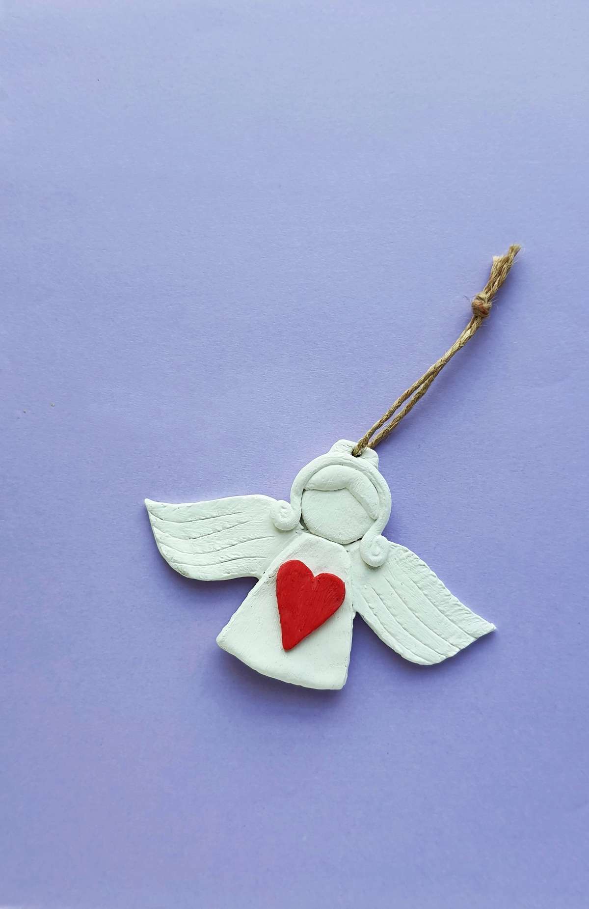 White clay angel Christmas tree ornament with sculpted wings and hair and red sculpted clay heart prominent in center.