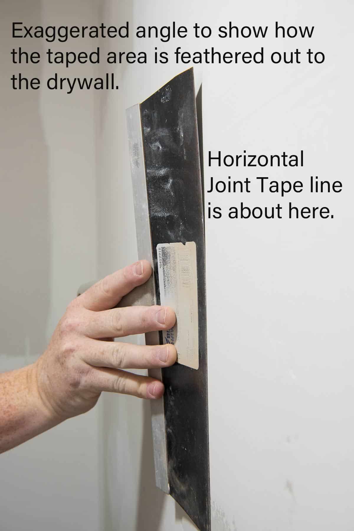 A hand holding a wide drywall knife to show how to feather joint compound over taped areas.