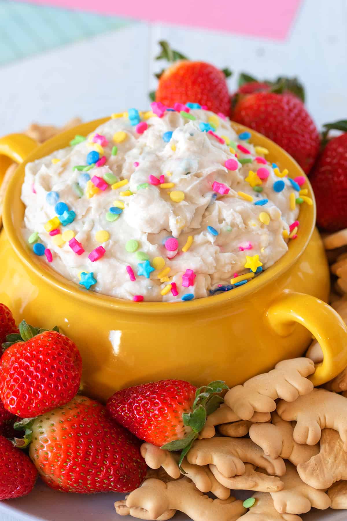 A bowl of cake batter dip with brightly colored sprinkles on top and strawberries and animal crackers around the bowl.