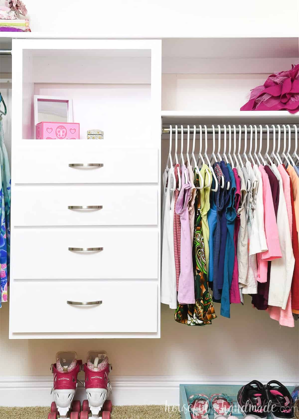 A pale pink closet with a white dresser and clothes hanging to the right of it.
