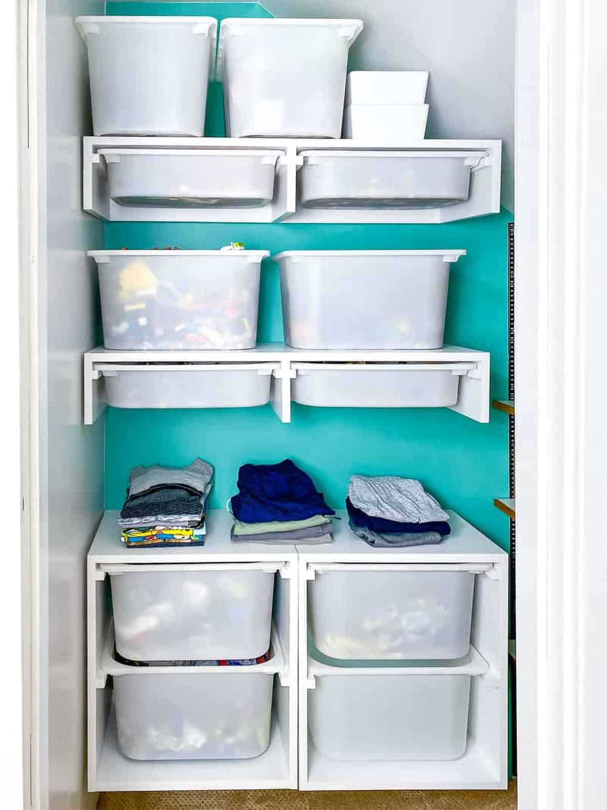 A white closet with a teal accent wall and white shelves filled with buckets of toys.