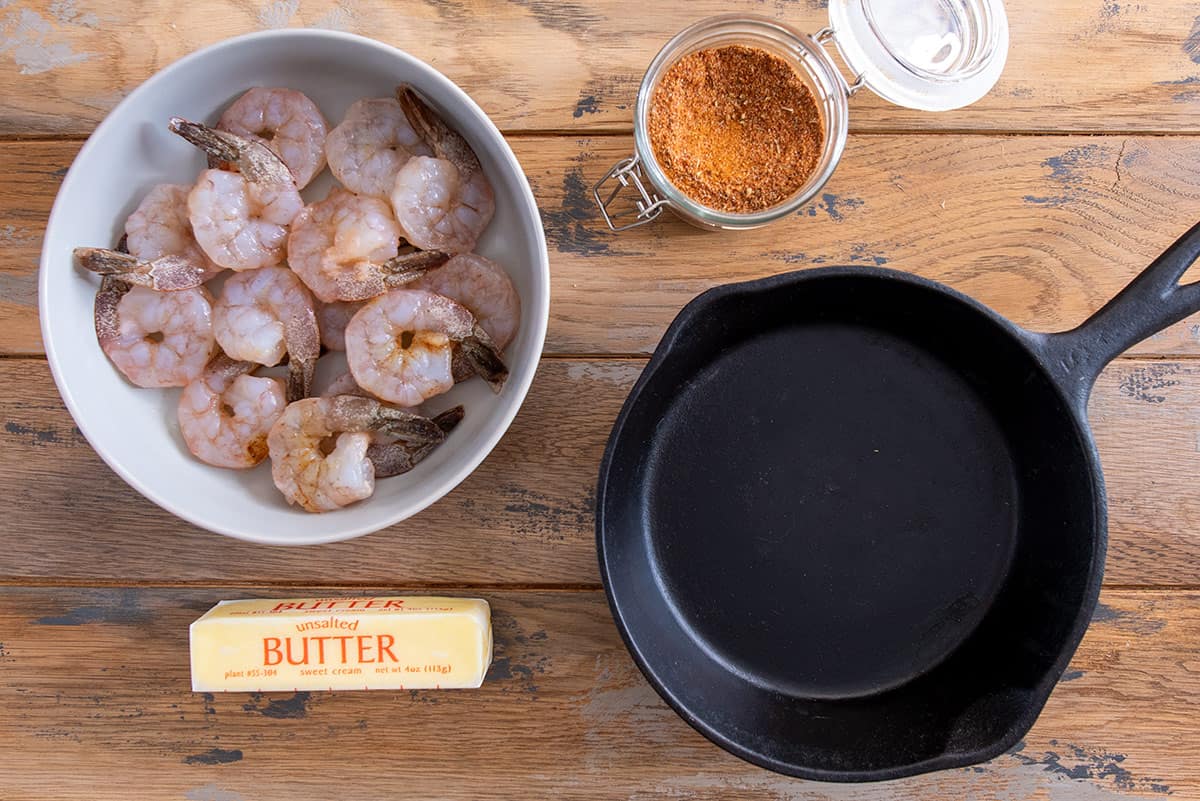 Ingredients for shrimp recipe including shrimp with tails on, Cajun seasoning blend, unsalted butter, and a cast iron pan.