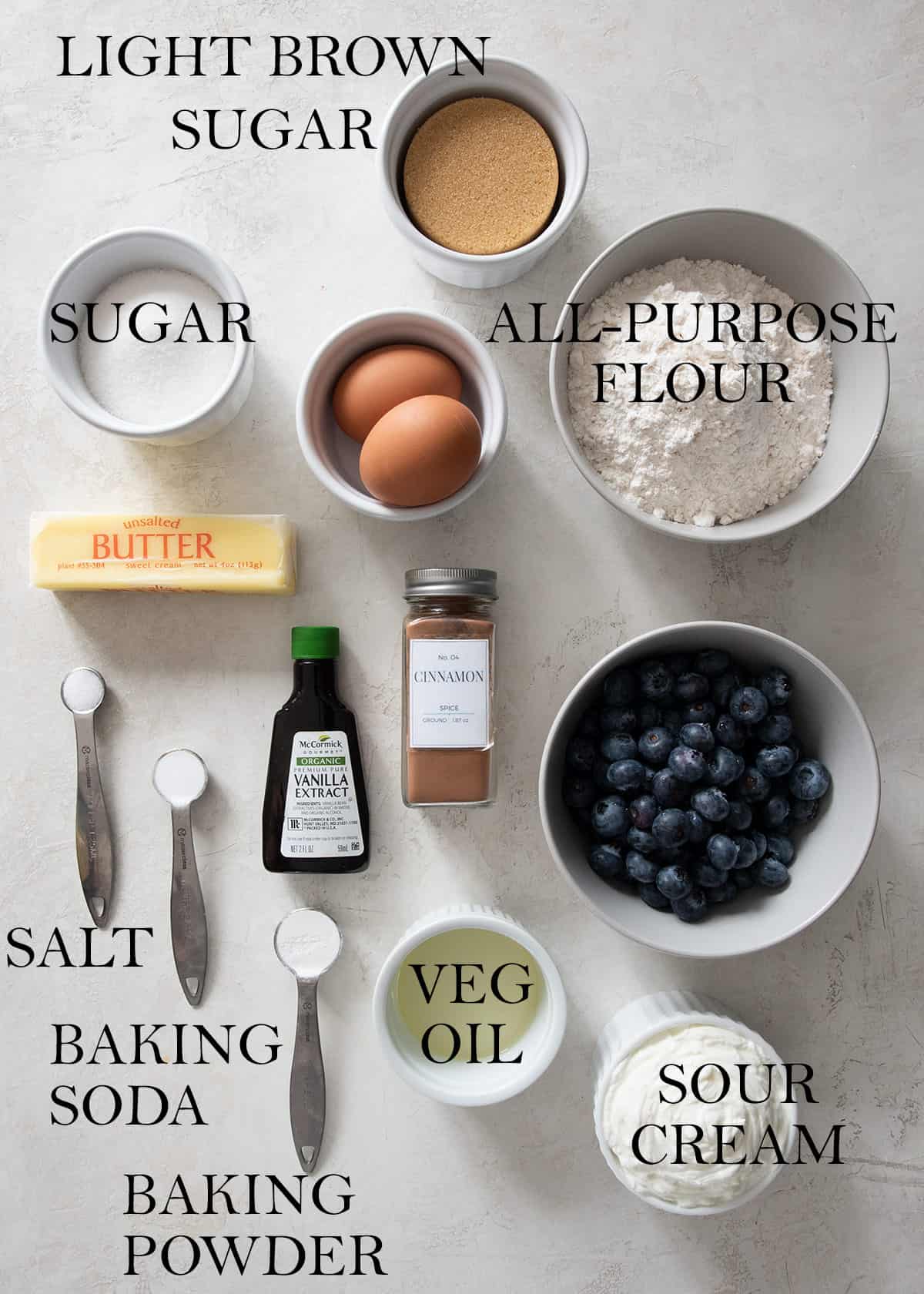 Ingredients for a blueberry coffee cake laid out on a table with text labels such as sugar, flour, unsalted, butter, and cinnamon.