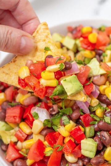 A hand holding a tortilla chip full of cowboy caviar with diced tomatoes, corn, beans, and avocado.