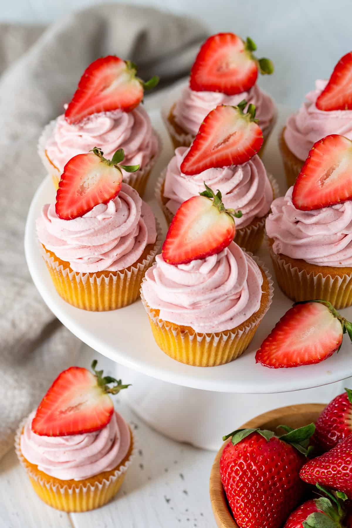 Strawberry Cream Cheese Frosted Cupcakes on white cake plate with sliced strawberries atop.