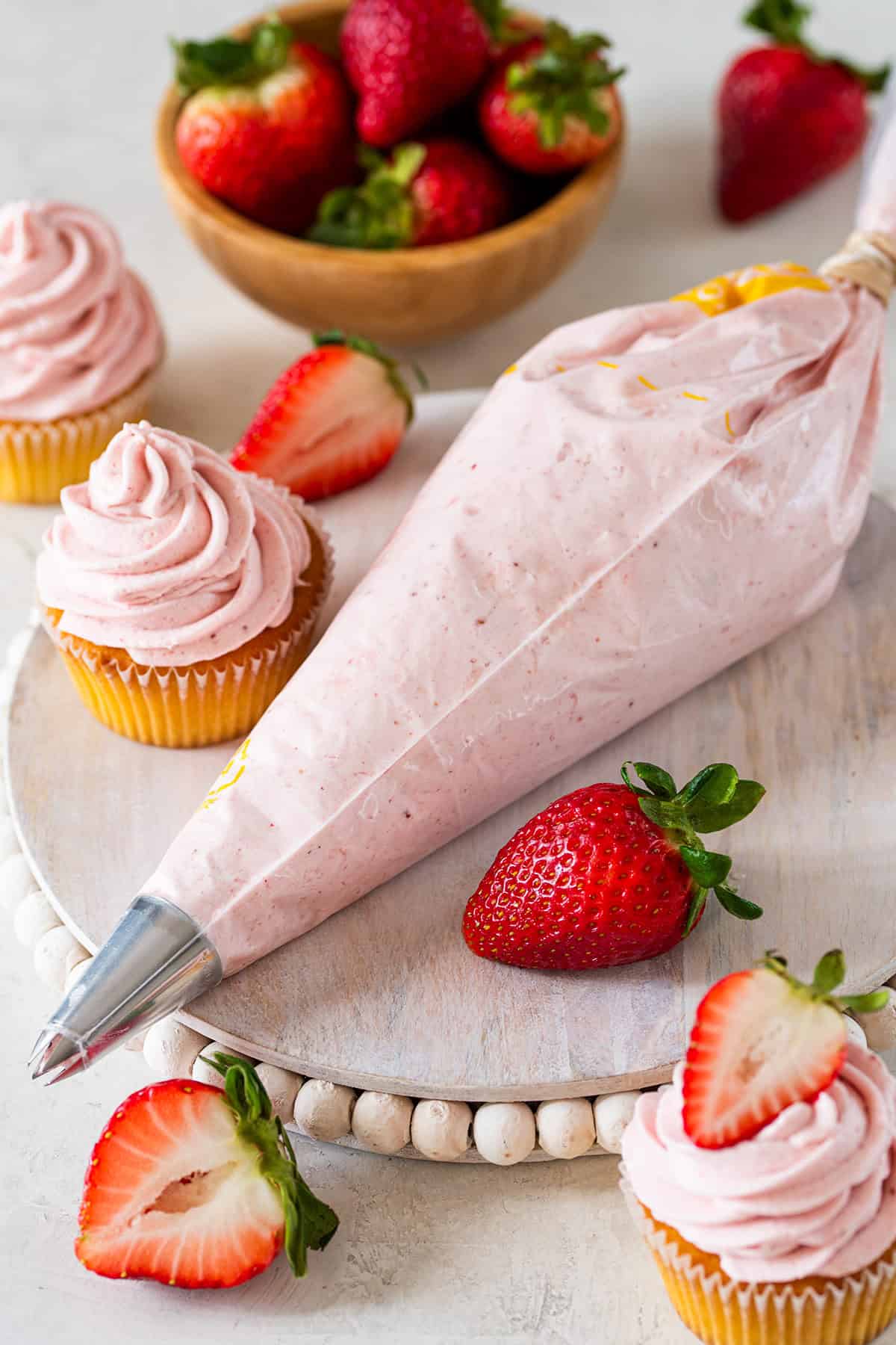 Piping bag full of Strawberry Cream Cheese icing on white cake plate with three iced cupcakes and a bowl of strawberries.