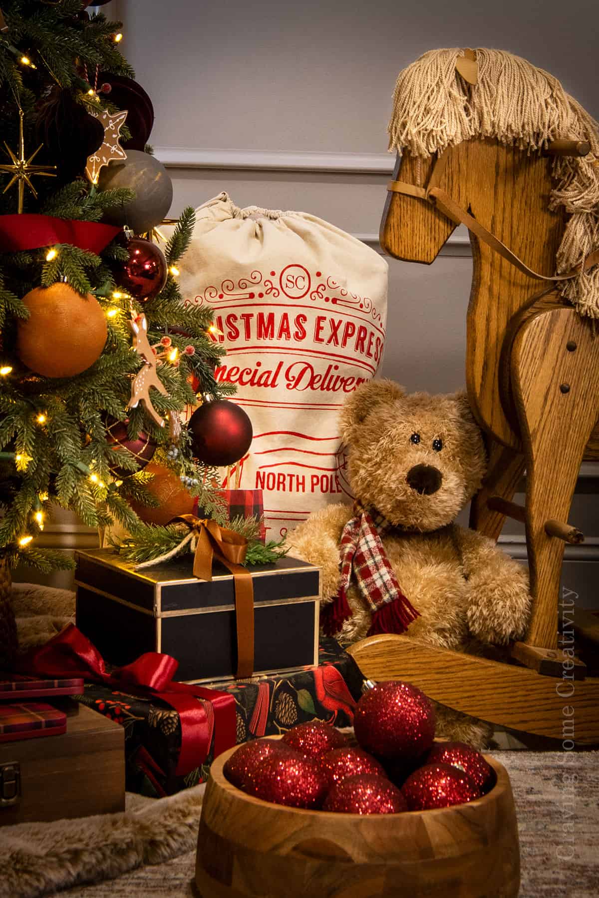 A vignette of antique rocking horse, teddy bear, Santa sack, ornaments, and gifts under a holiday tree.