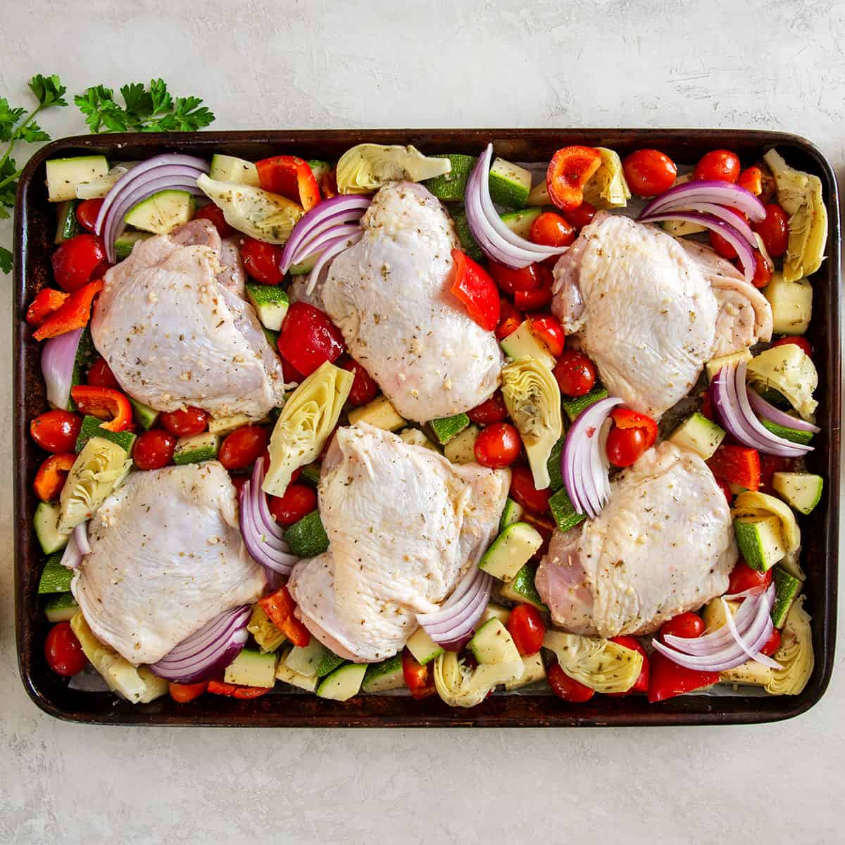 A rimmed baking sheet with chicken thighs and vegetables laid out before baking.