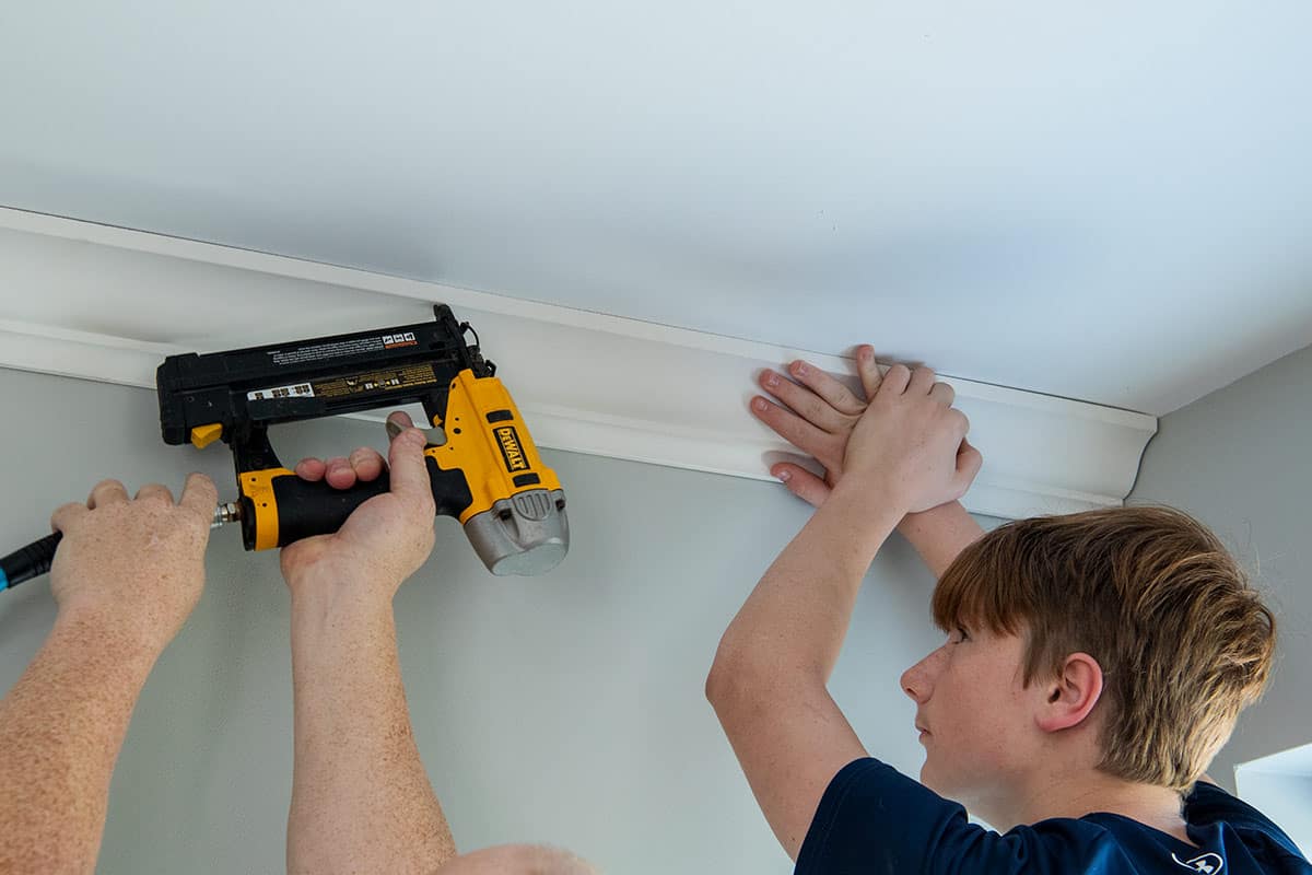 A teenage boy holding crown molding in place while another male nails it in.