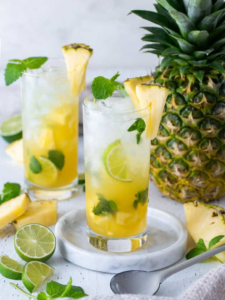 Two Pineapple Mojito Cocktails in tall glasses with limes and pineapples as garnish.